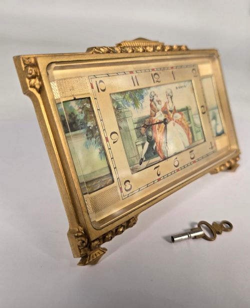 fig 1. A rectangular gilt brass Art Deco strut clock featuring an illustrated courtly scene on the central dial surrounded by numeric markers.