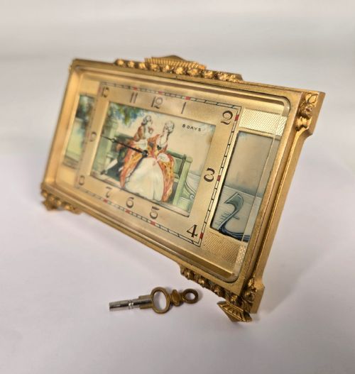 fig 2. A rectangular gilt brass Art Deco strut clock featuring an illustrated courtly scene on the central dial surrounded by numeric markers.