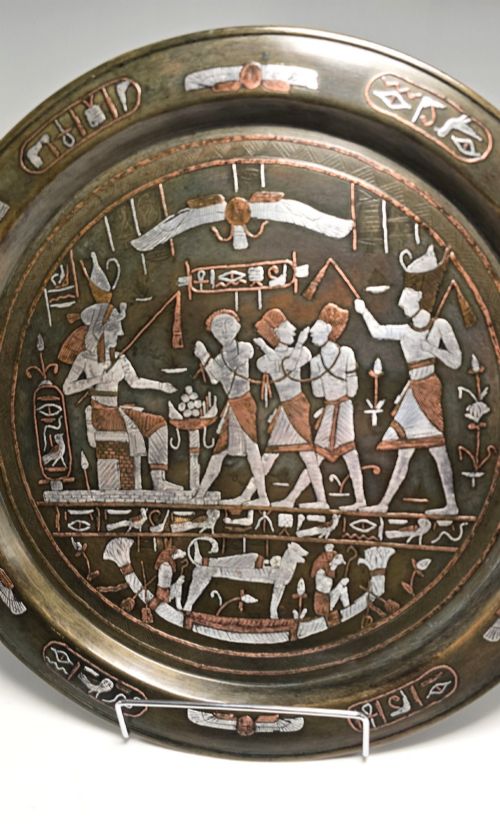 fig 1. A circular bronze charger decorated with silver and copper inlays depicting pharaonic figures, hieroglyphs, and a winged sun deity.