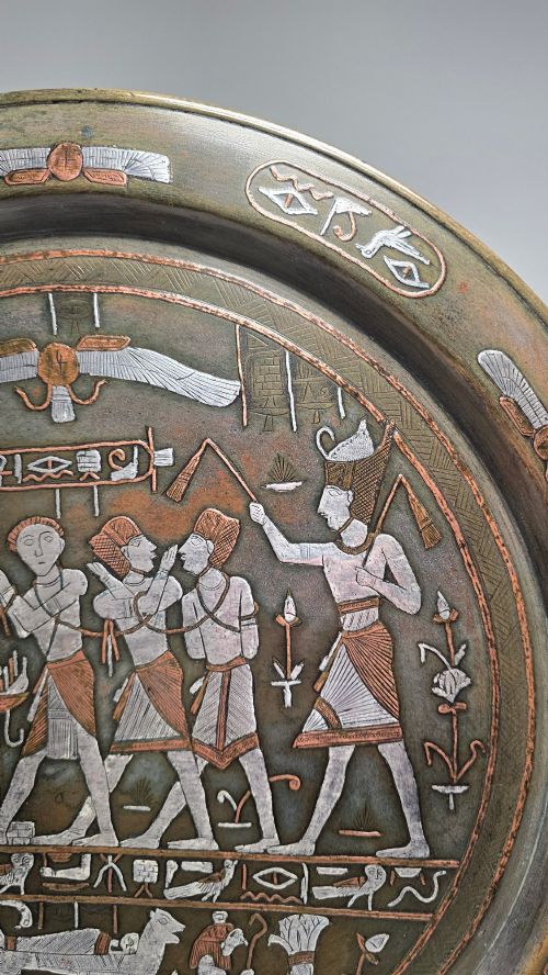 fig 2. A circular bronze charger decorated with silver and copper inlays depicting pharaonic figures, hieroglyphs, and a winged sun deity.