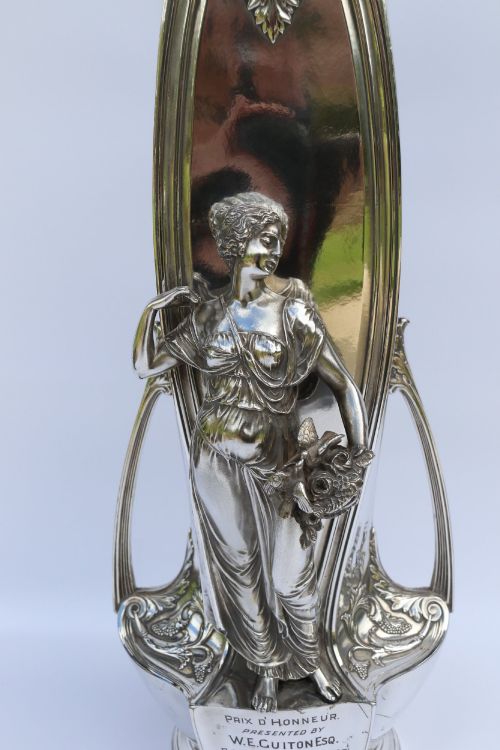 fig 3. A tall silver-plated Art Nouveau vase featuring a relief figure of a maiden holding flowers, twin handles, an engraved presentation inscription at the base, and a clear glass liner.