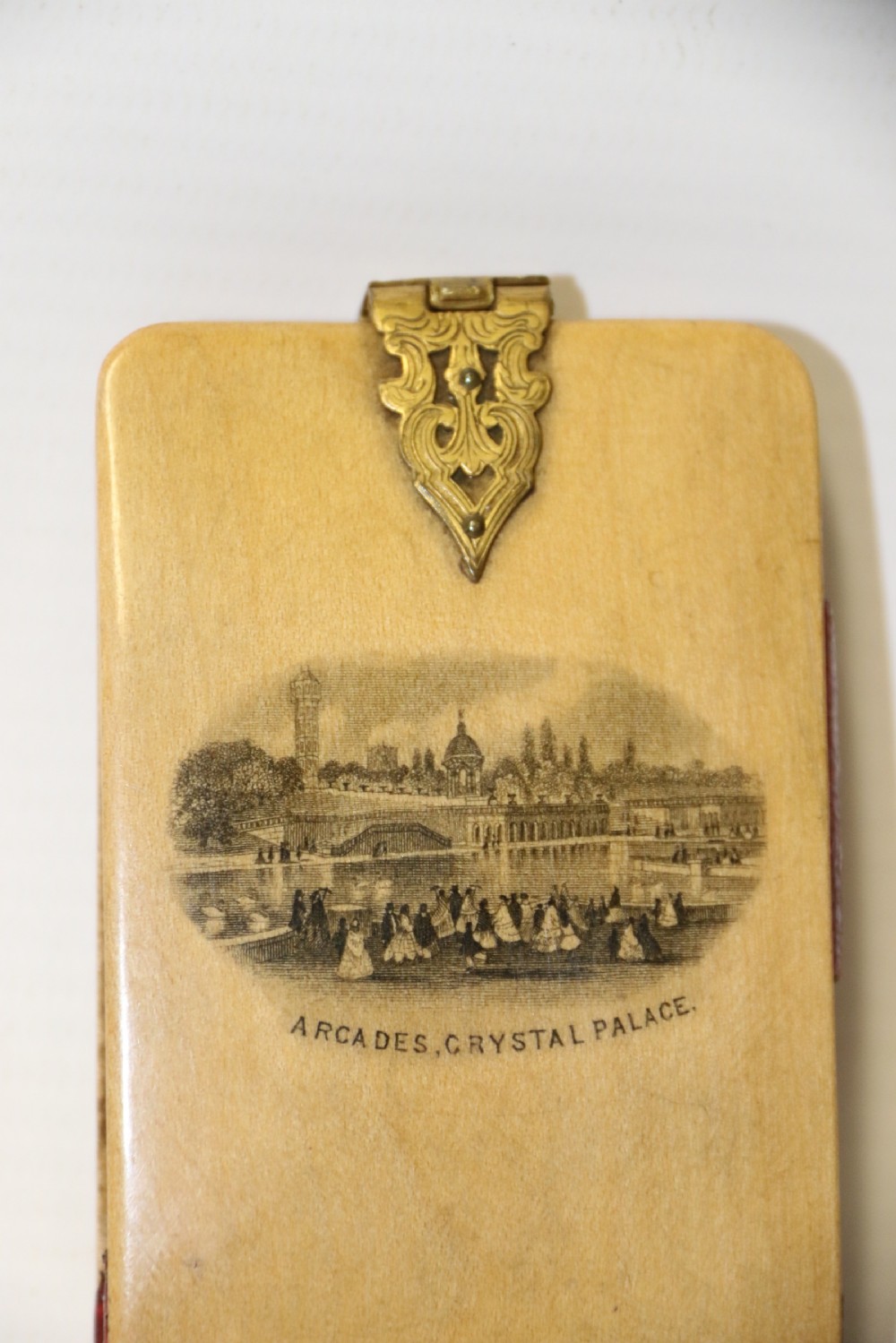 An antique Victorian Mauchline ware wooden pocket notebook displaying a black oval transfer print depicting the Arcades at Crystal Palace, featuring an ornate brass top clasp. View II