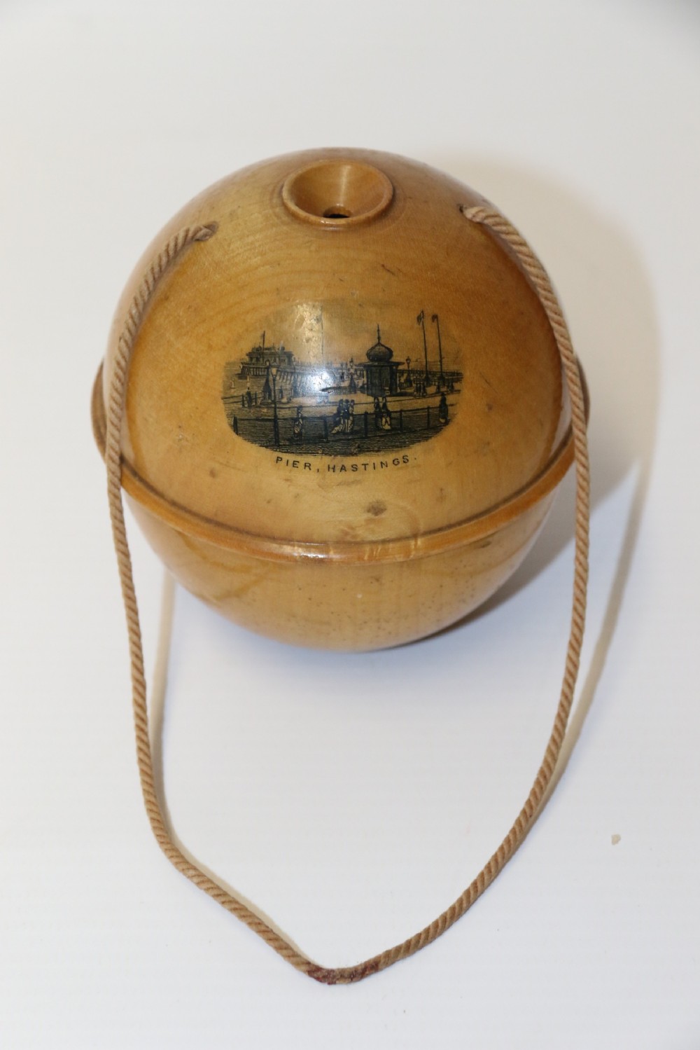 A spherical turned wooden Mauchline ware string holder featuring a black transfer print of Hastings Pier and a hanging cord.