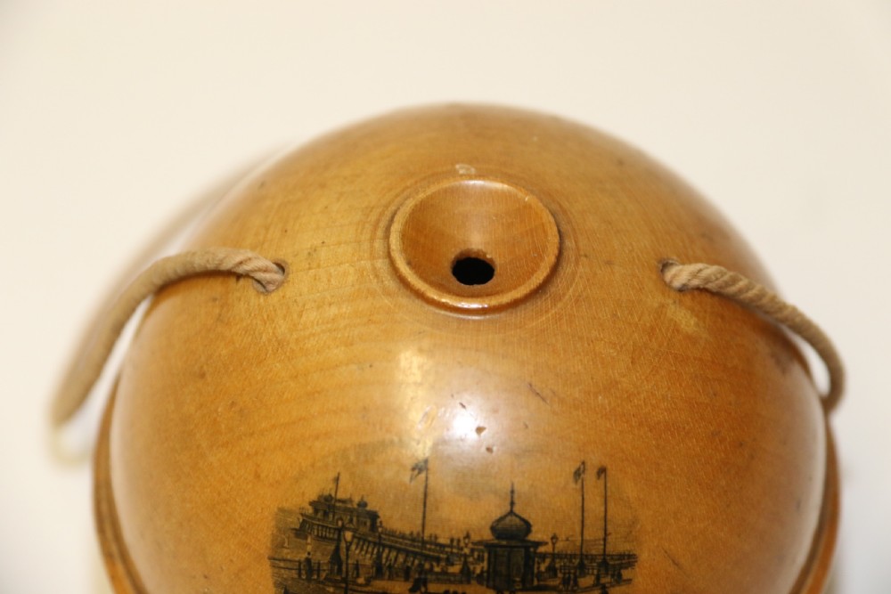 A spherical turned wooden Mauchline ware string holder featuring a black transfer print of Hastings Pier and a hanging cord. View V