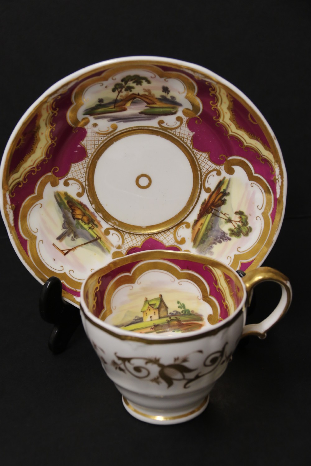 An early 19th-century English porcelain cabinet cup and saucer decorated with magenta borders, gold scrollwork gilding, and hand-painted miniature landscape vignettes. View II