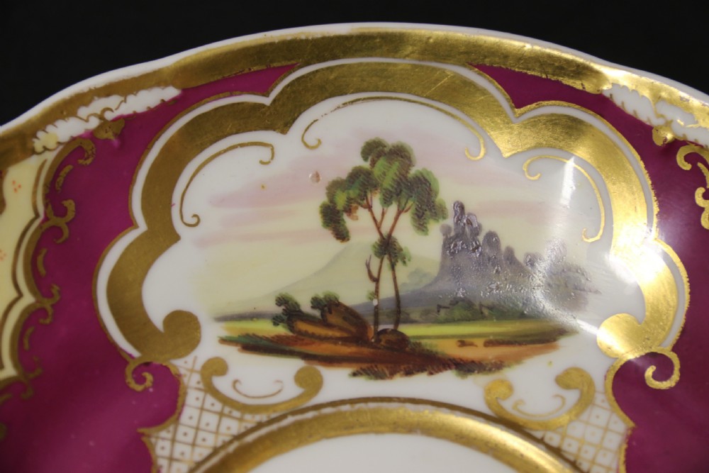 An early 19th-century English porcelain cabinet cup and saucer decorated with magenta borders, gold scrollwork gilding, and hand-painted miniature landscape vignettes. View V