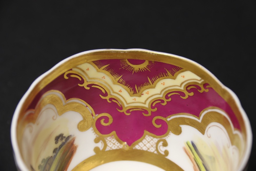 An early 19th-century English porcelain cabinet cup and saucer decorated with magenta borders, gold scrollwork gilding, and hand-painted miniature landscape vignettes. View XI