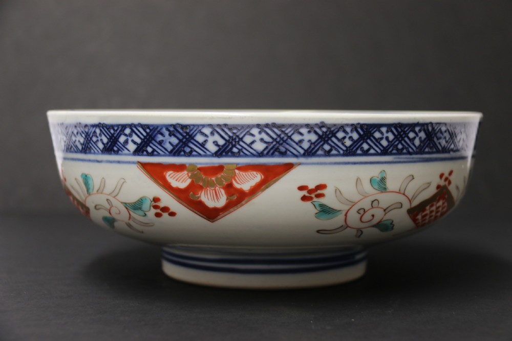 A round hand-painted Japanese porcelain bowl featuring underglaze blue geometric rim borders, iron-red floral panel motifs, and gilded details on a white background. View XIII