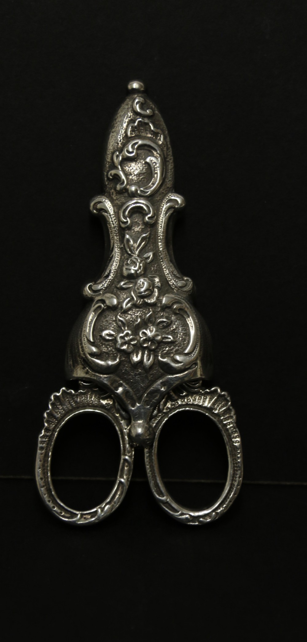 A mid-19th-century silver scissor sheath featuring embossed floral scrollwork details holding a pair of silver scissors with oval finger loops.