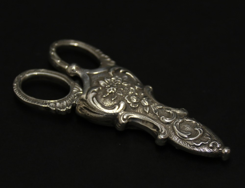 A mid-19th-century silver scissor sheath featuring embossed floral scrollwork details holding a pair of silver scissors with oval finger loops. View II