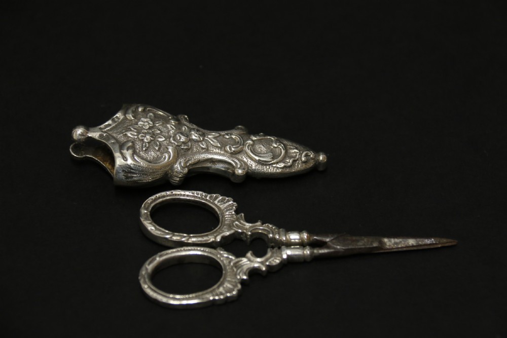 A mid-19th-century silver scissor sheath featuring embossed floral scrollwork details holding a pair of silver scissors with oval finger loops. View IV