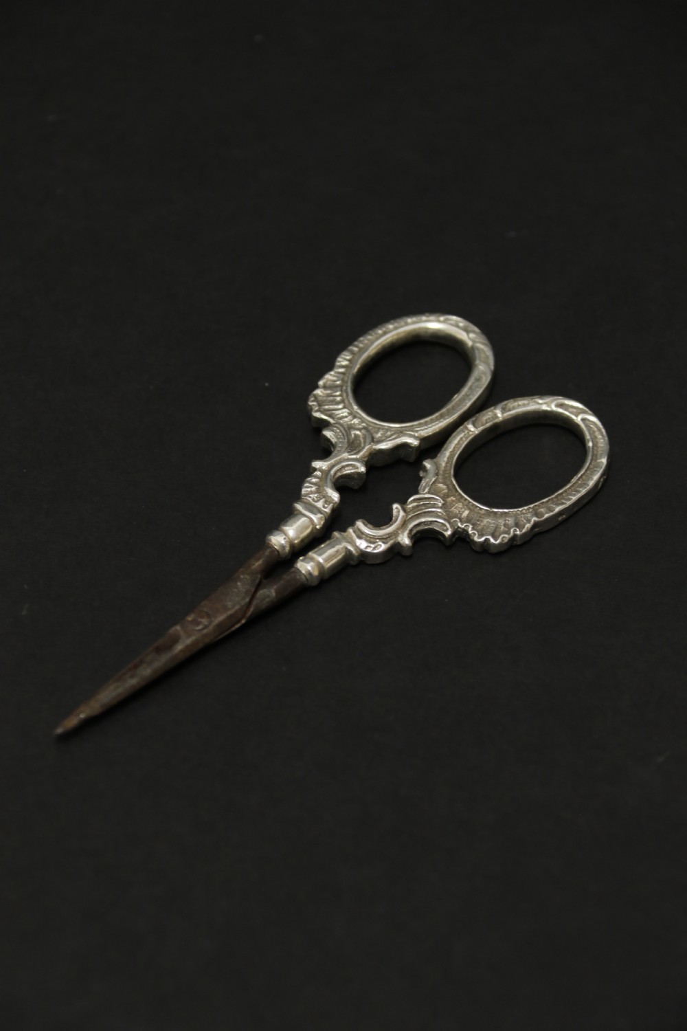 A mid-19th-century silver scissor sheath featuring embossed floral scrollwork details holding a pair of silver scissors with oval finger loops. View V