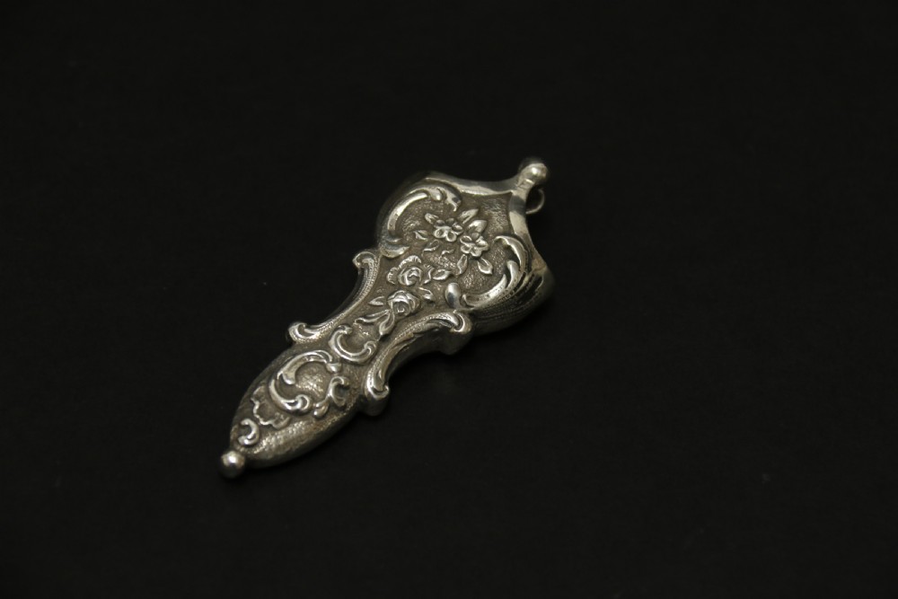A mid-19th-century silver scissor sheath featuring embossed floral scrollwork details holding a pair of silver scissors with oval finger loops. View VI