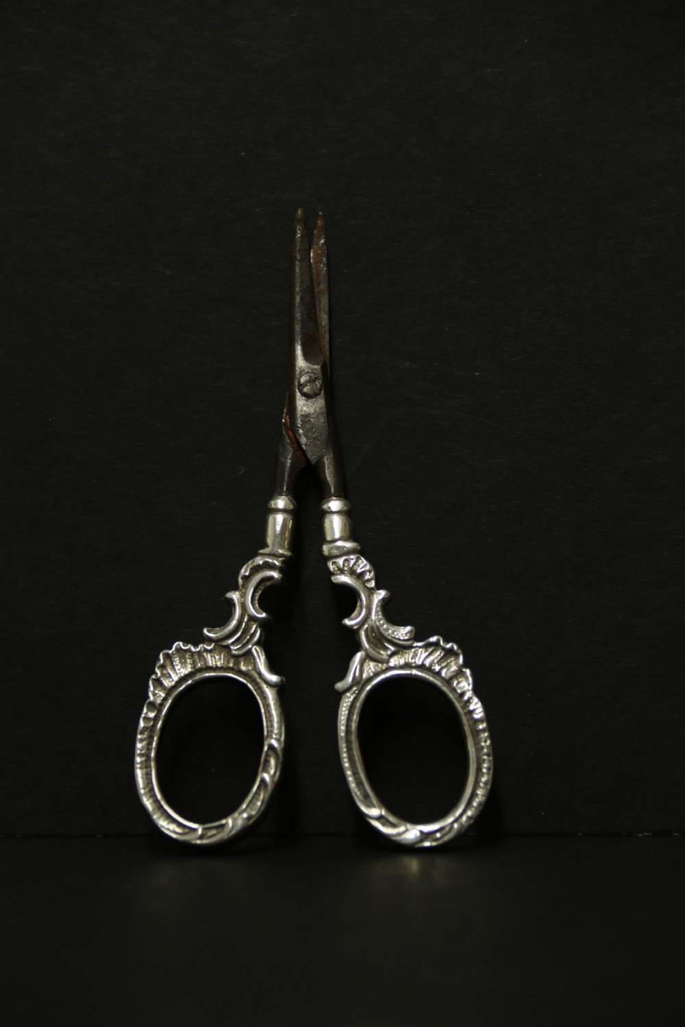 A mid-19th-century silver scissor sheath featuring embossed floral scrollwork details holding a pair of silver scissors with oval finger loops. View VII