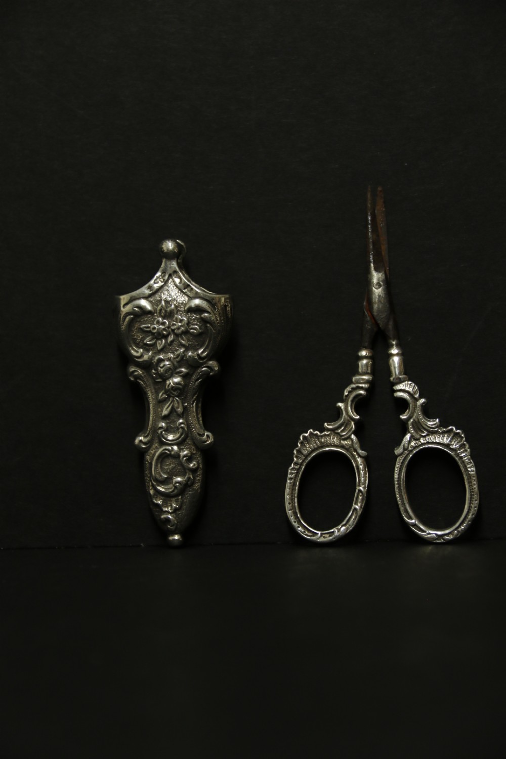 A mid-19th-century silver scissor sheath featuring embossed floral scrollwork details holding a pair of silver scissors with oval finger loops. View VIII