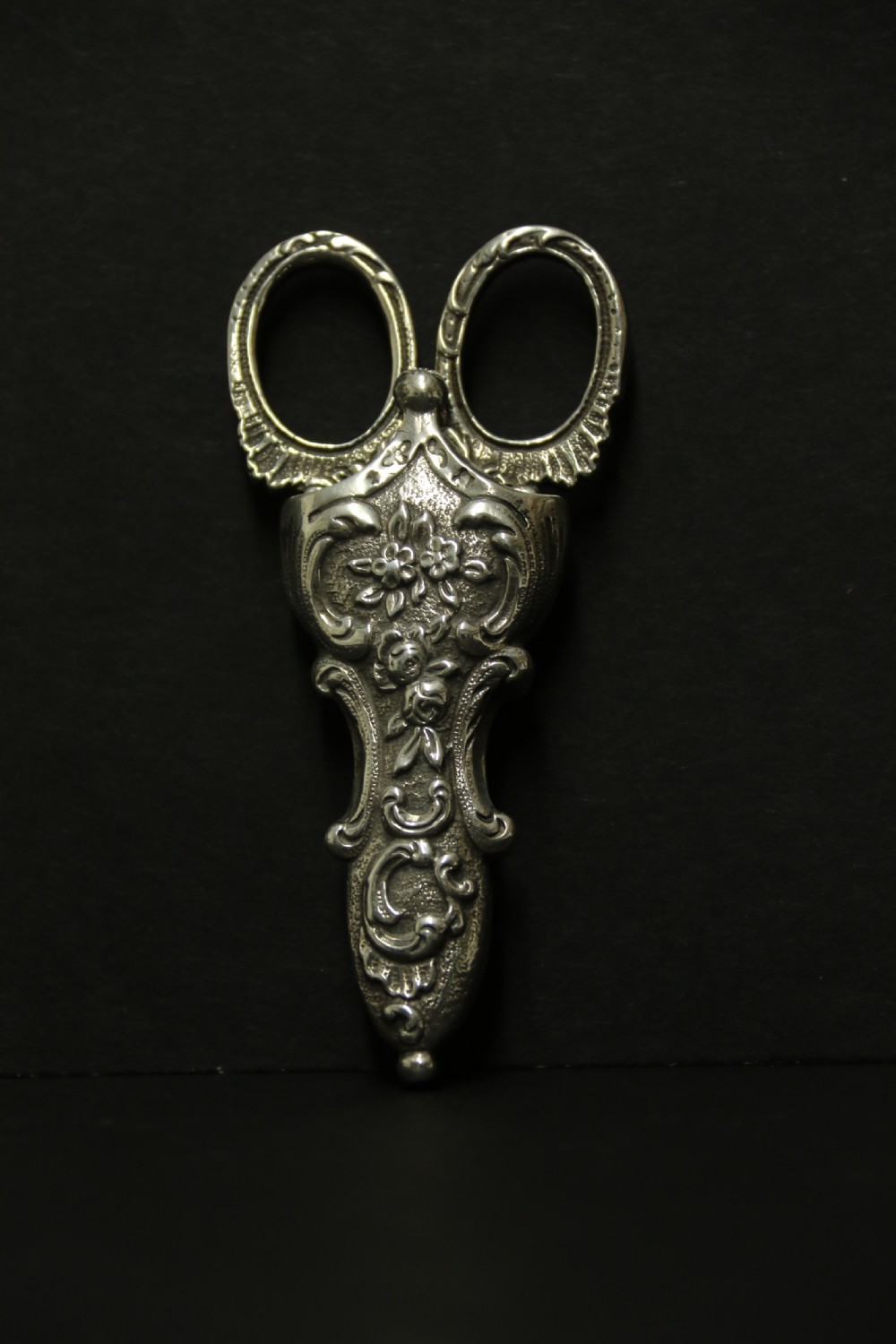 A mid-19th-century silver scissor sheath featuring embossed floral scrollwork details holding a pair of silver scissors with oval finger loops. View IX