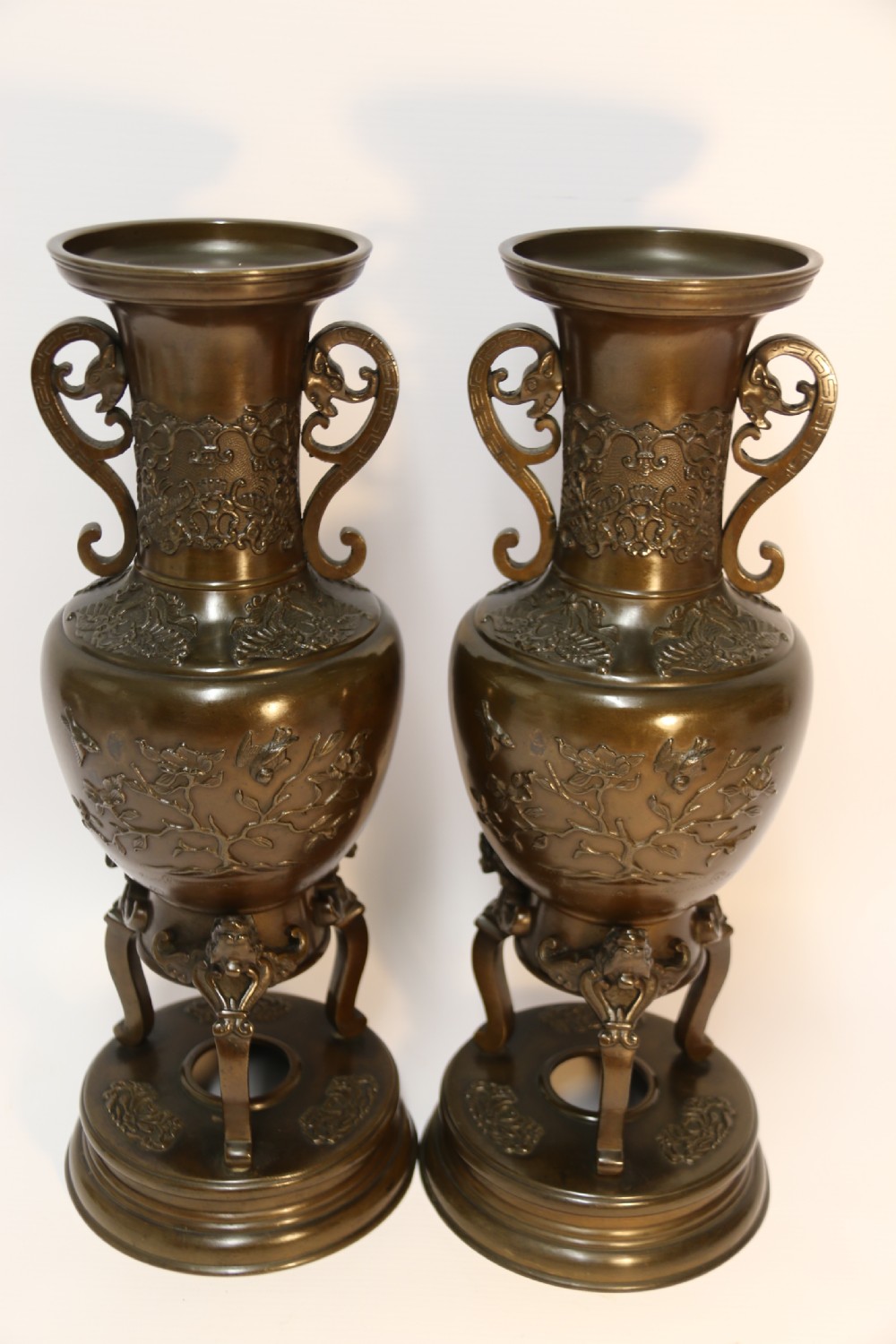 A pair of patinated Japanese Meiji period bronze vases featuring low-relief floral carving, curved scroll handles, and tripod legs resting on circular bases.