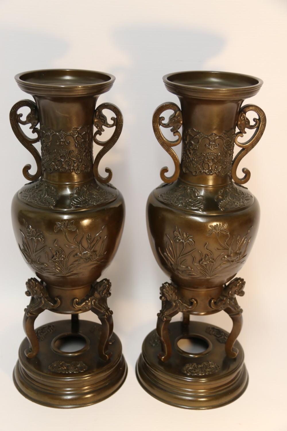 A pair of patinated Japanese Meiji period bronze vases featuring low-relief floral carving, curved scroll handles, and tripod legs resting on circular bases. View II