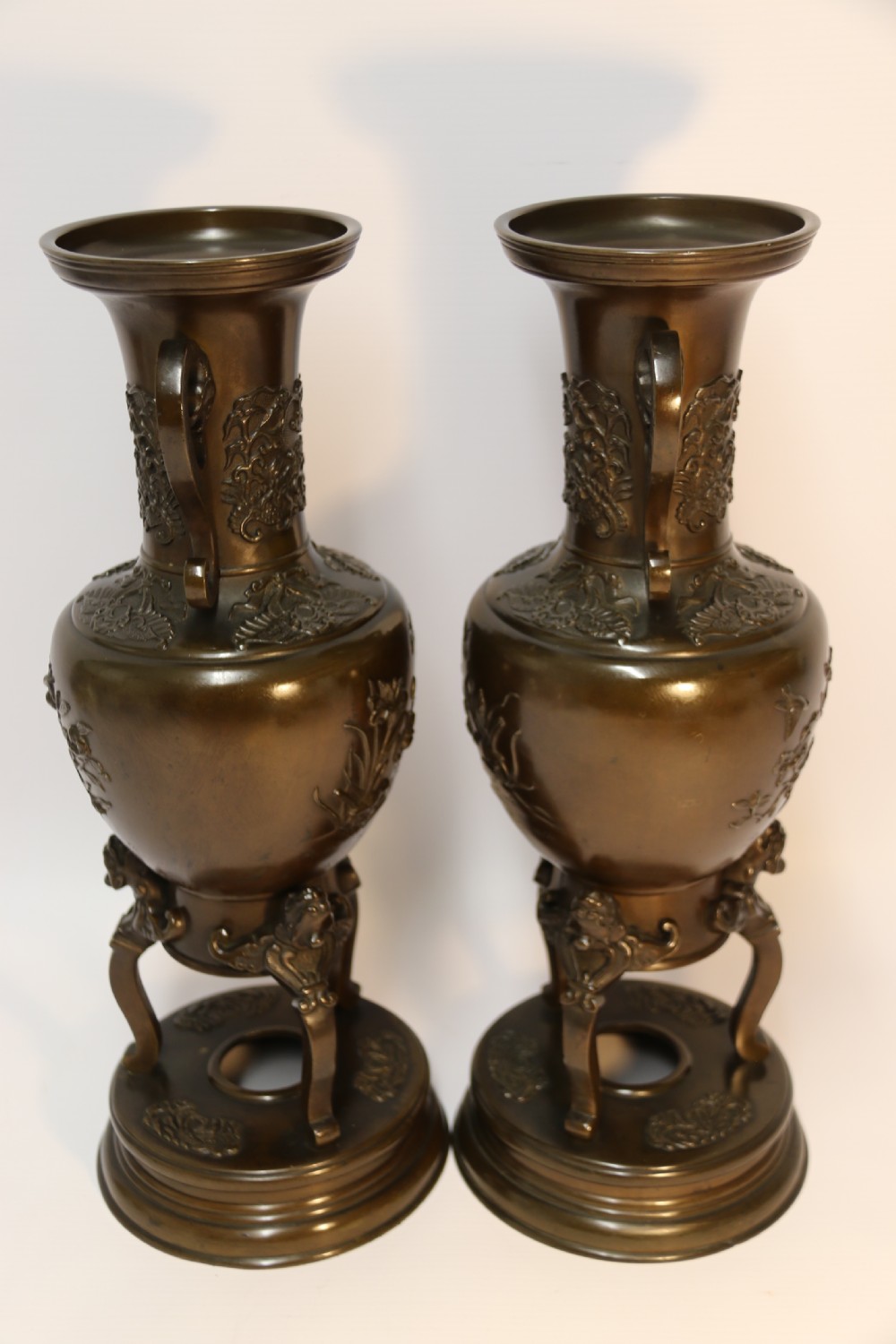 A pair of patinated Japanese Meiji period bronze vases featuring low-relief floral carving, curved scroll handles, and tripod legs resting on circular bases. View III