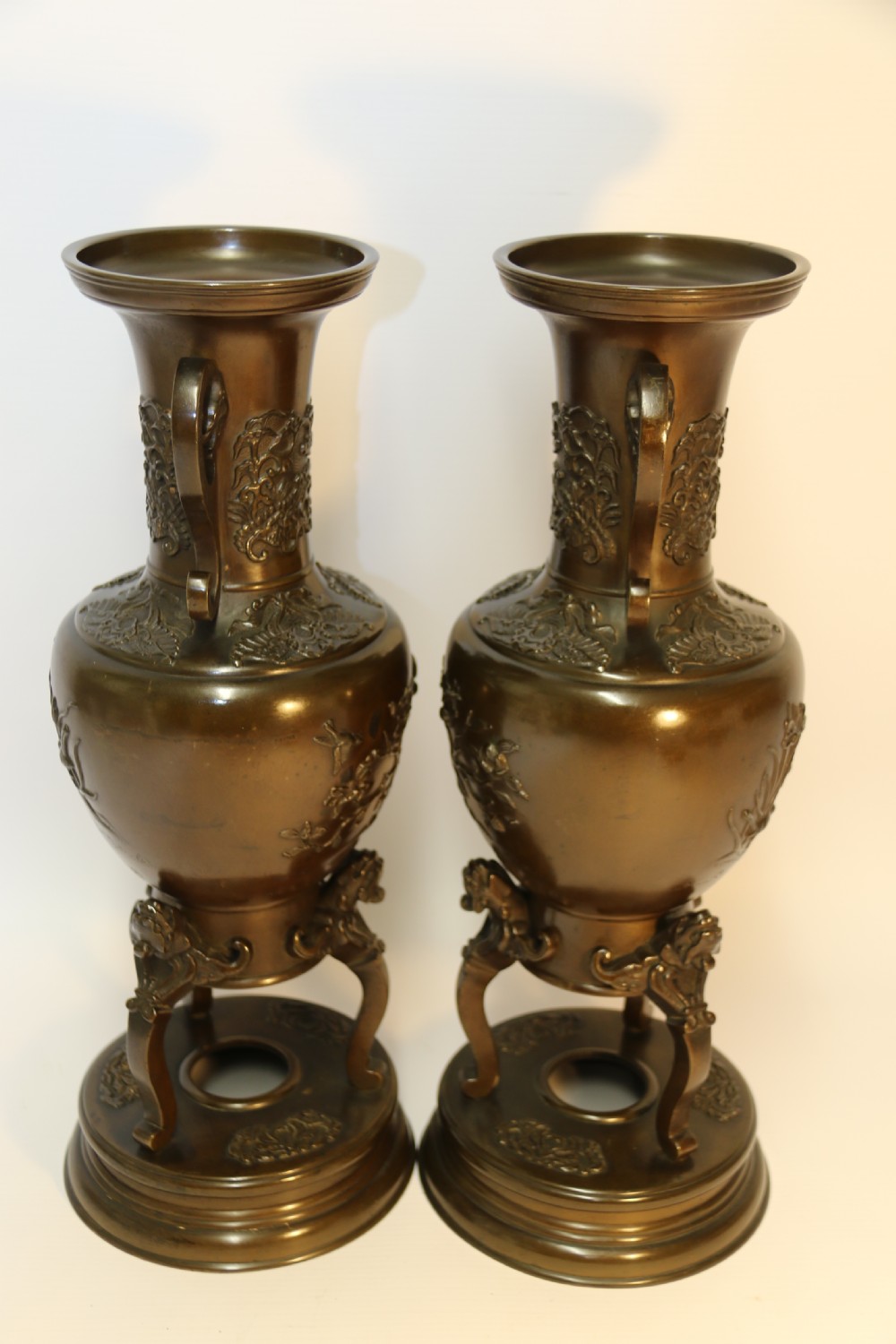 A pair of patinated Japanese Meiji period bronze vases featuring low-relief floral carving, curved scroll handles, and tripod legs resting on circular bases. View IV