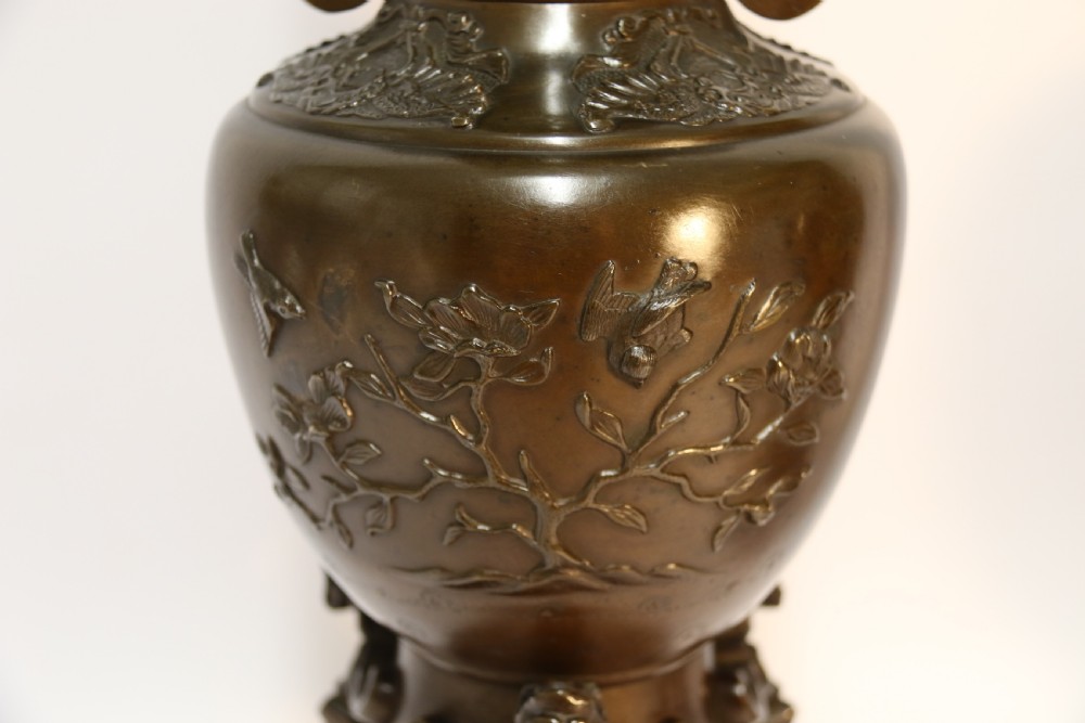 A pair of patinated Japanese Meiji period bronze vases featuring low-relief floral carving, curved scroll handles, and tripod legs resting on circular bases. View V
