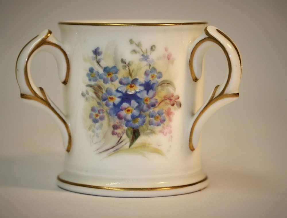 A white miniature porcelain loving cup featuring twin gilded scroll handles and a hand-painted blue floral spray.