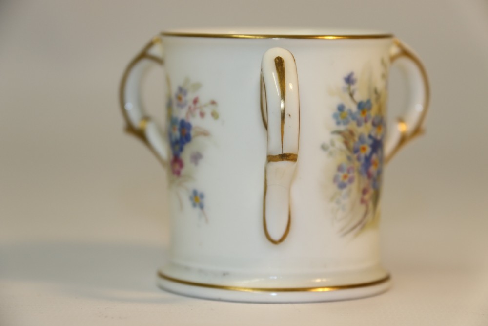 A white miniature porcelain loving cup featuring twin gilded scroll handles and a hand-painted blue floral spray. View II