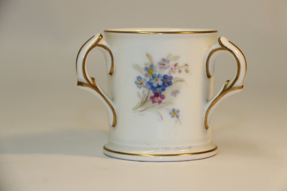 A white miniature porcelain loving cup featuring twin gilded scroll handles and a hand-painted blue floral spray. View III