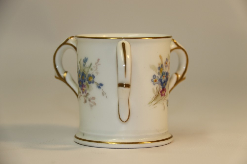 A white miniature porcelain loving cup featuring twin gilded scroll handles and a hand-painted blue floral spray. View IV