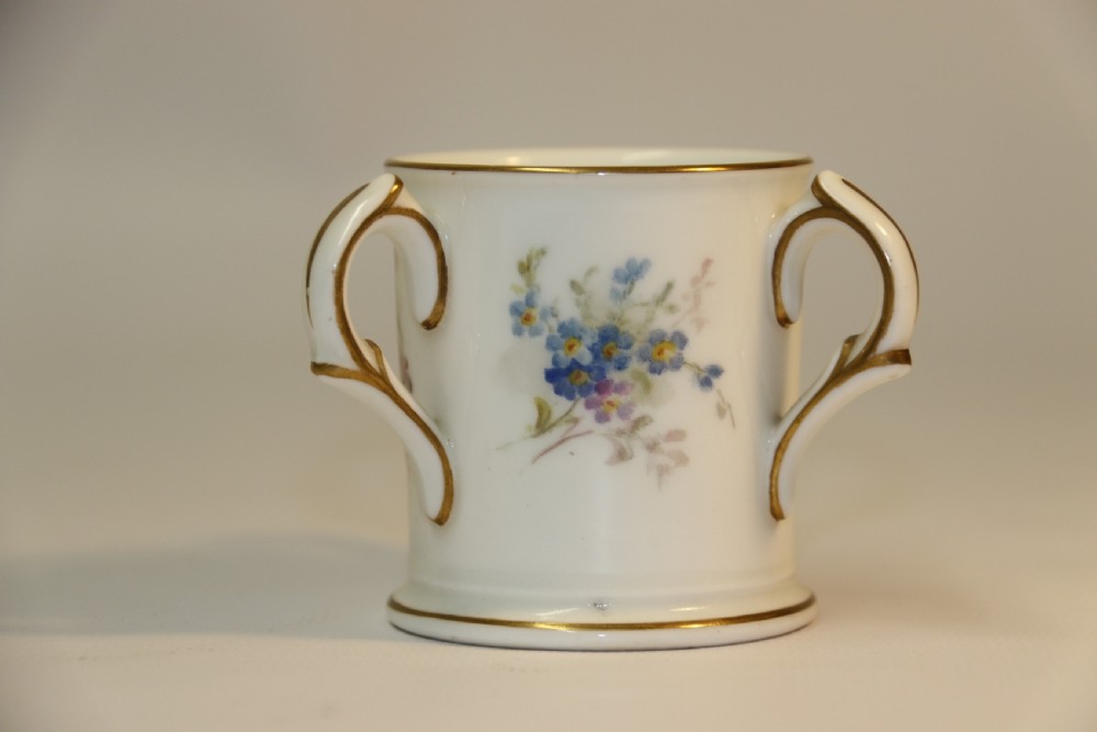 A white miniature porcelain loving cup featuring twin gilded scroll handles and a hand-painted blue floral spray. View V