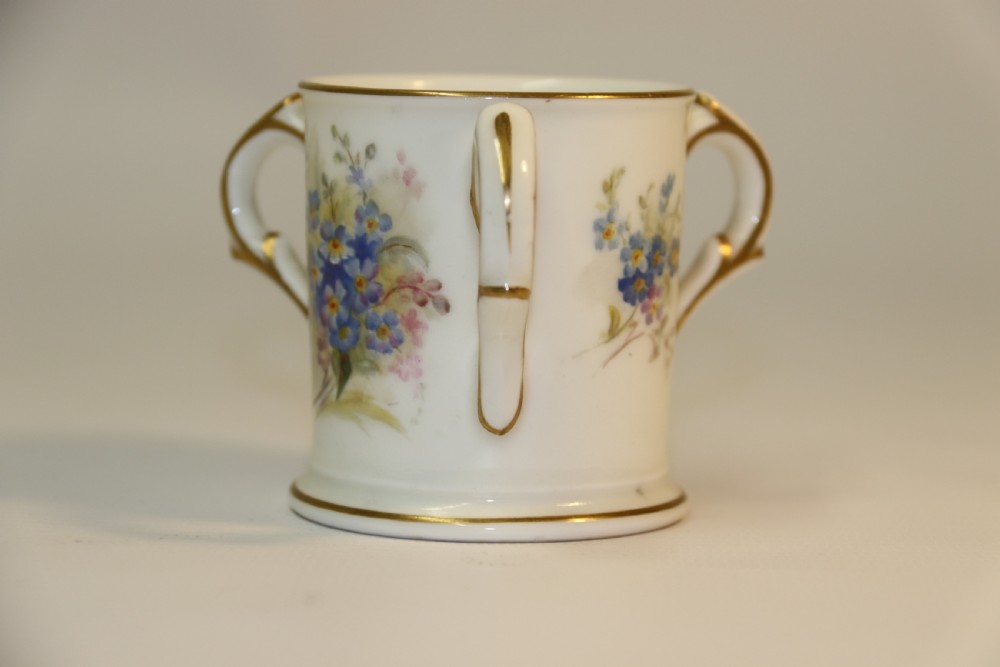 A white miniature porcelain loving cup featuring twin gilded scroll handles and a hand-painted blue floral spray. View VII