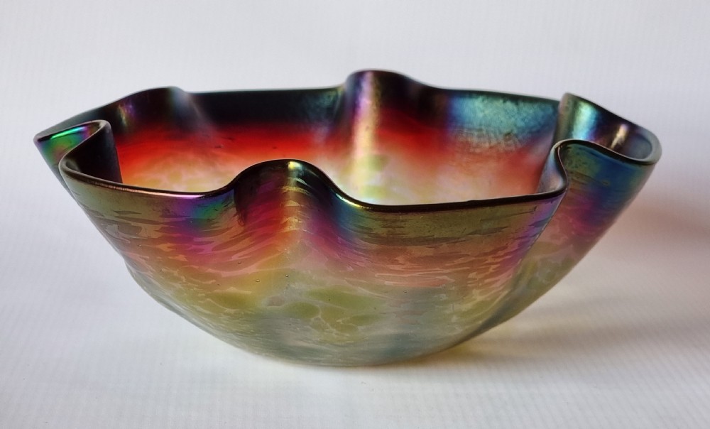 A hand-blown glass bowl featuring an undulating ruffled rim and a shimmering iridescent finish in shades of green, purple, and red.
