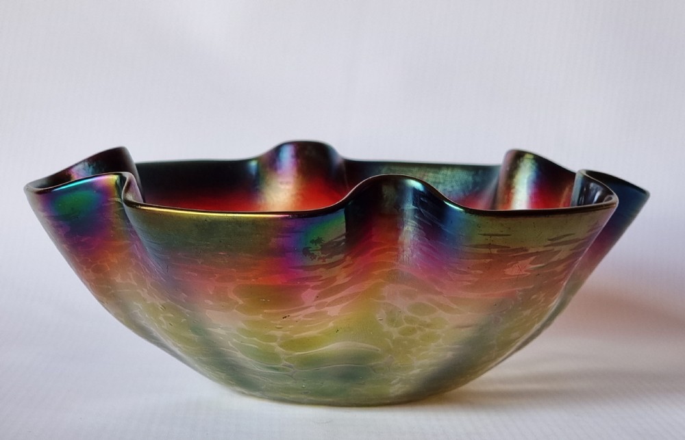 A hand-blown glass bowl featuring an undulating ruffled rim and a shimmering iridescent finish in shades of green, purple, and red. View IX