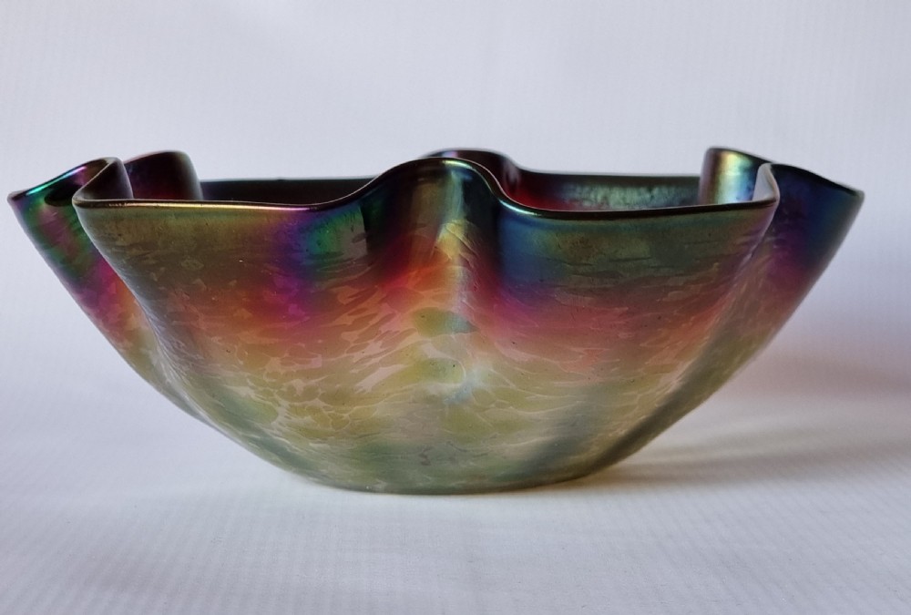 A hand-blown glass bowl featuring an undulating ruffled rim and a shimmering iridescent finish in shades of green, purple, and red. View X