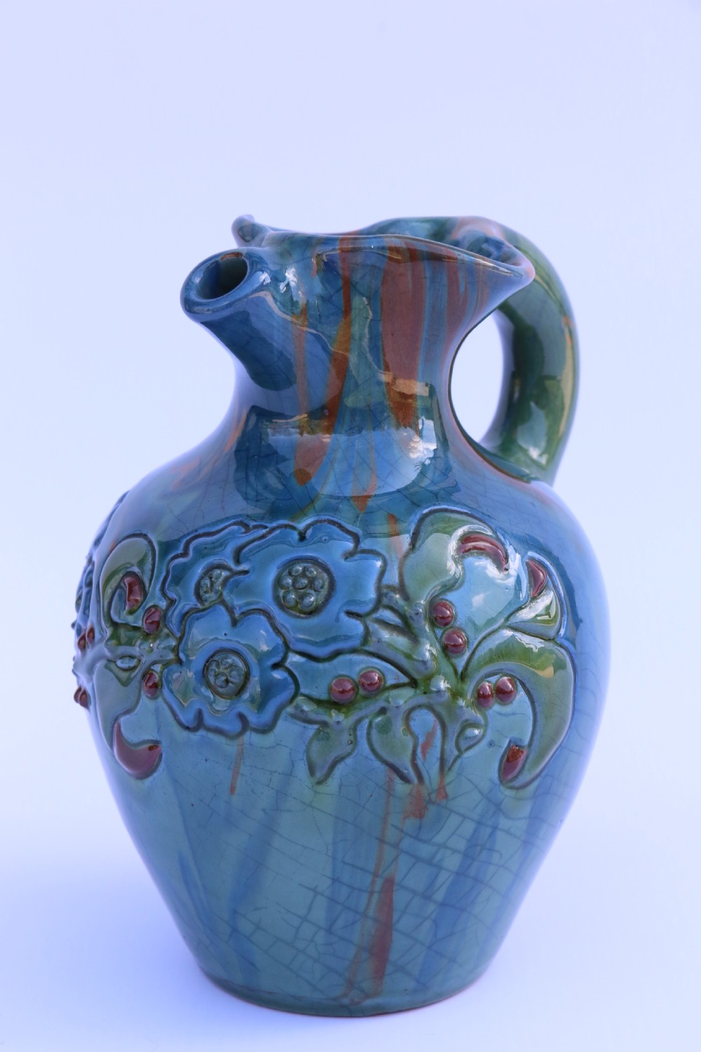 A teal blue Arts and Crafts earthenware studio pottery jug featuring raised floral slipwork motifs, red glazed highlights, and a pinched spout.
