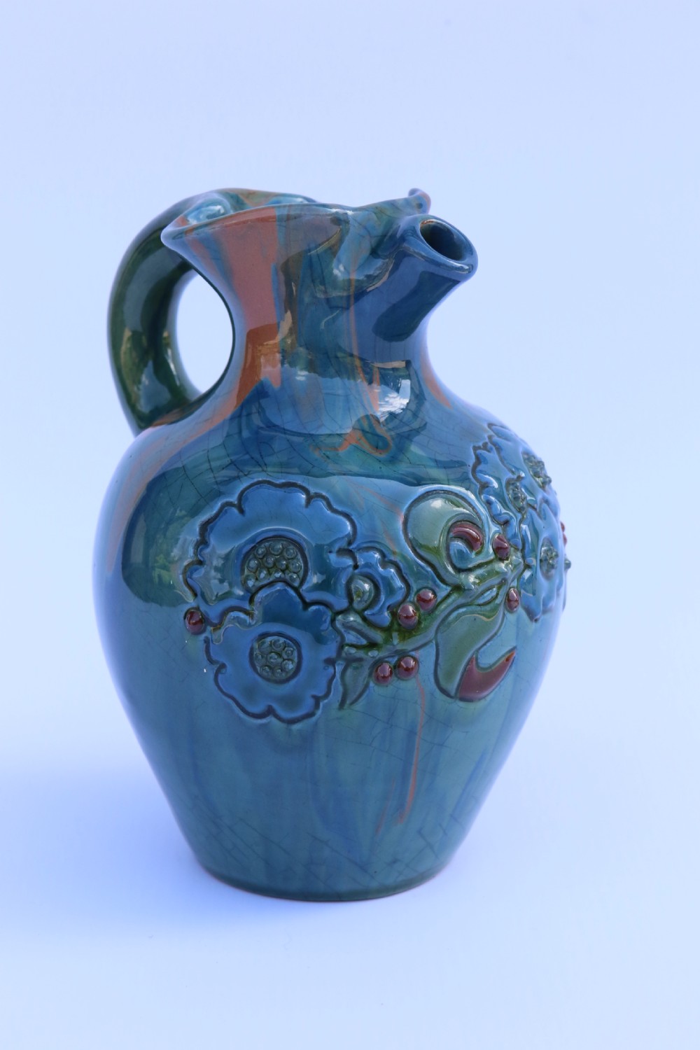 A teal blue Arts and Crafts earthenware studio pottery jug featuring raised floral slipwork motifs, red glazed highlights, and a pinched spout. View II