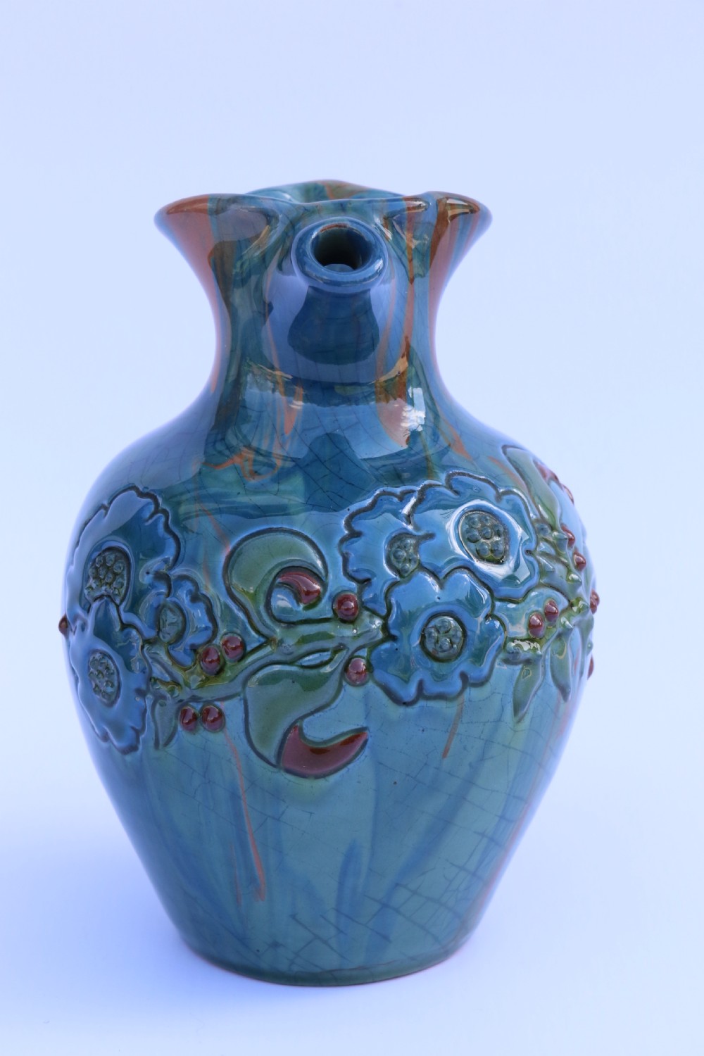 A teal blue Arts and Crafts earthenware studio pottery jug featuring raised floral slipwork motifs, red glazed highlights, and a pinched spout. View III