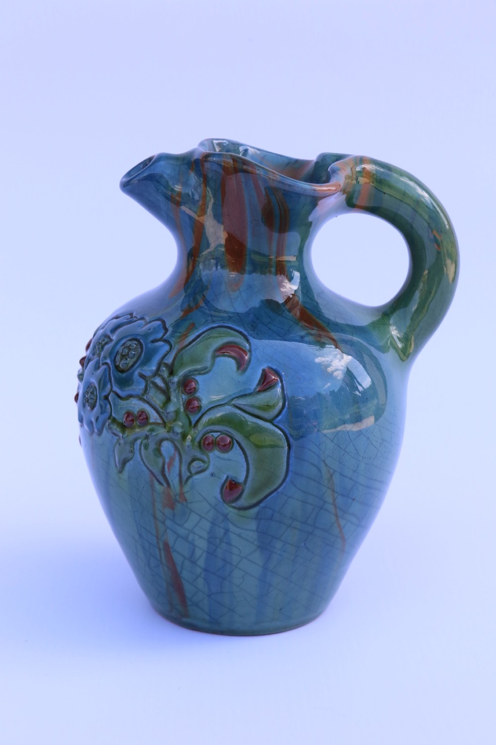 A teal blue Arts and Crafts earthenware studio pottery jug featuring raised floral slipwork motifs, red glazed highlights, and a pinched spout. View V