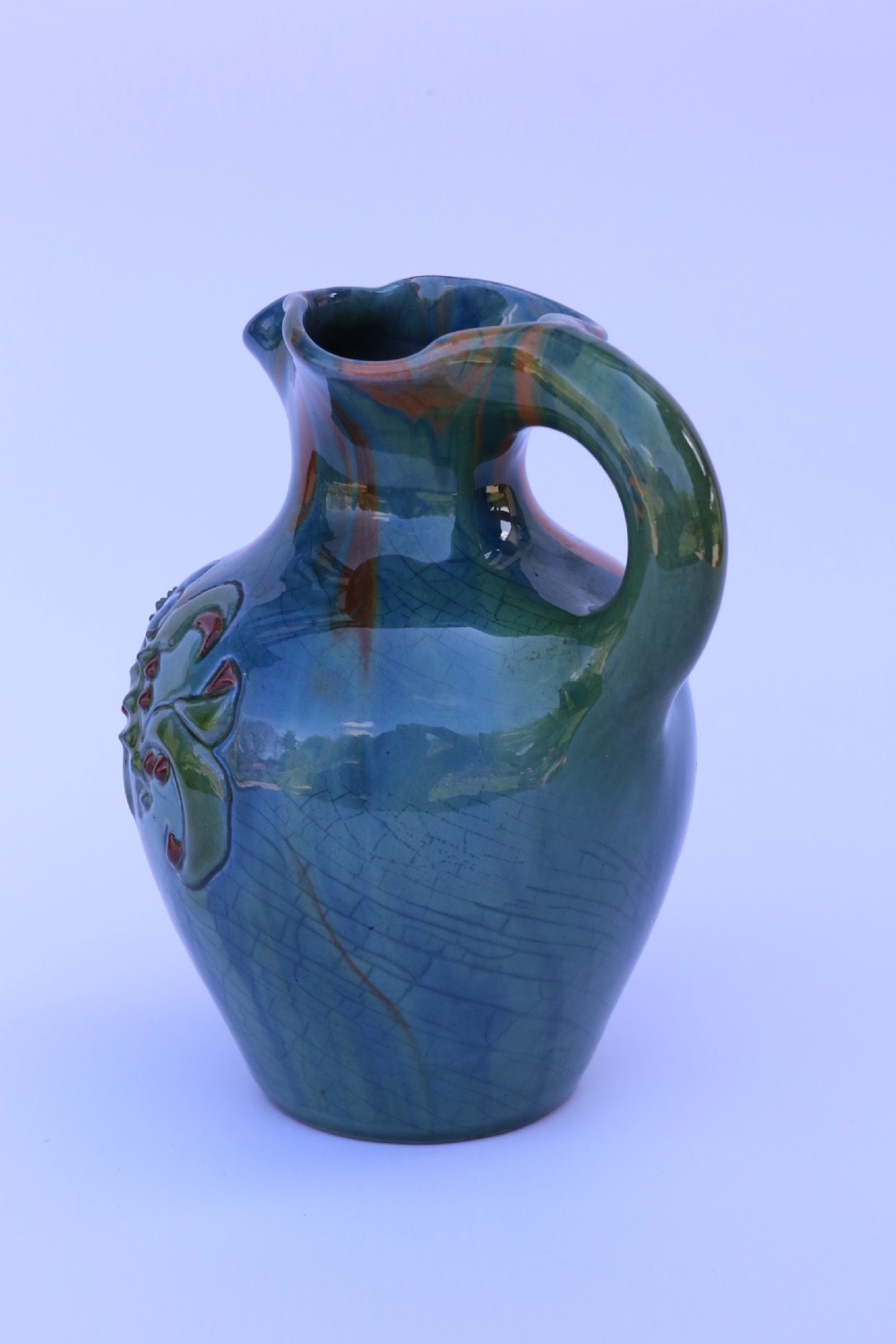 A teal blue Arts and Crafts earthenware studio pottery jug featuring raised floral slipwork motifs, red glazed highlights, and a pinched spout. View VII