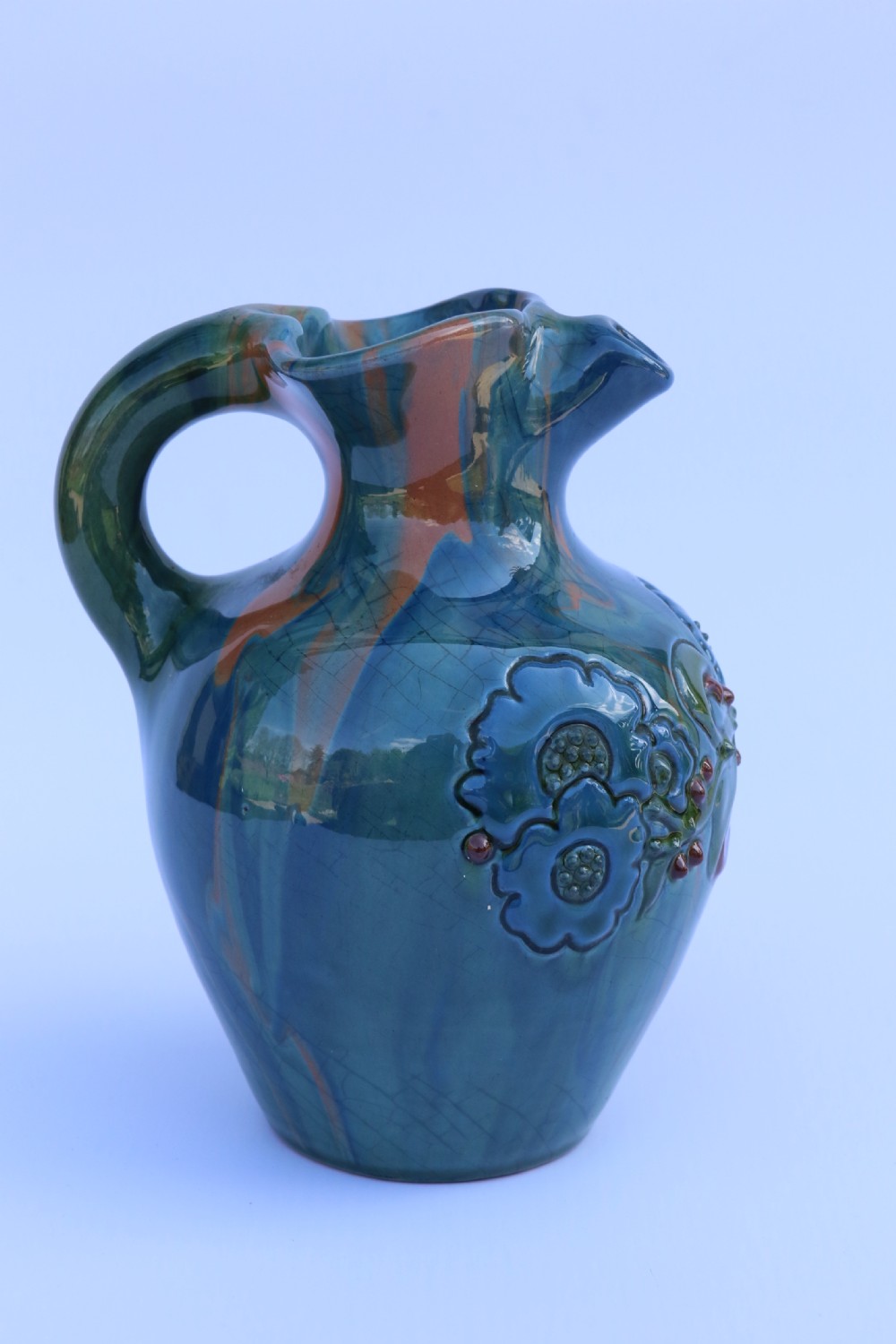 A teal blue Arts and Crafts earthenware studio pottery jug featuring raised floral slipwork motifs, red glazed highlights, and a pinched spout. View IX