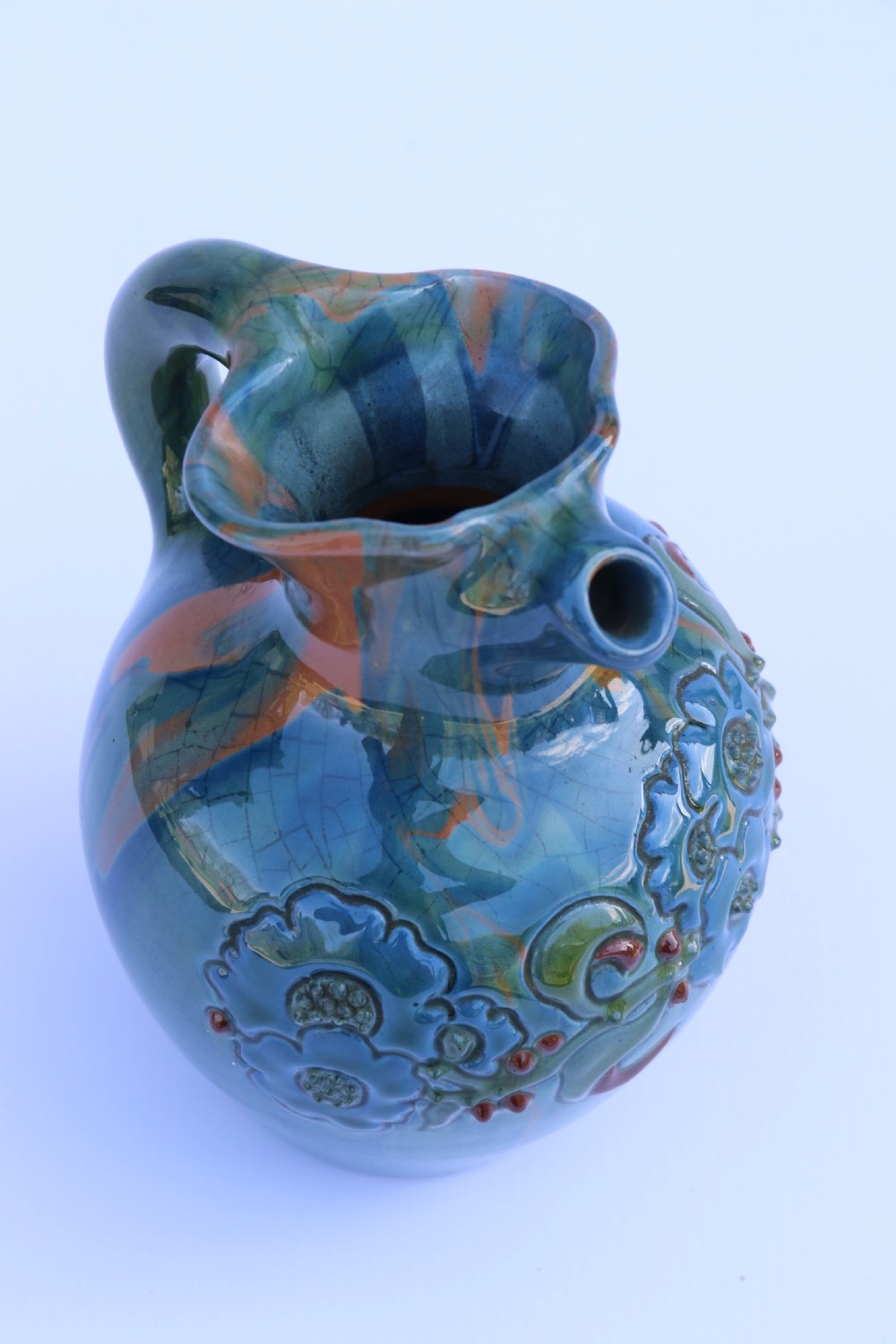 A teal blue Arts and Crafts earthenware studio pottery jug featuring raised floral slipwork motifs, red glazed highlights, and a pinched spout. View XI