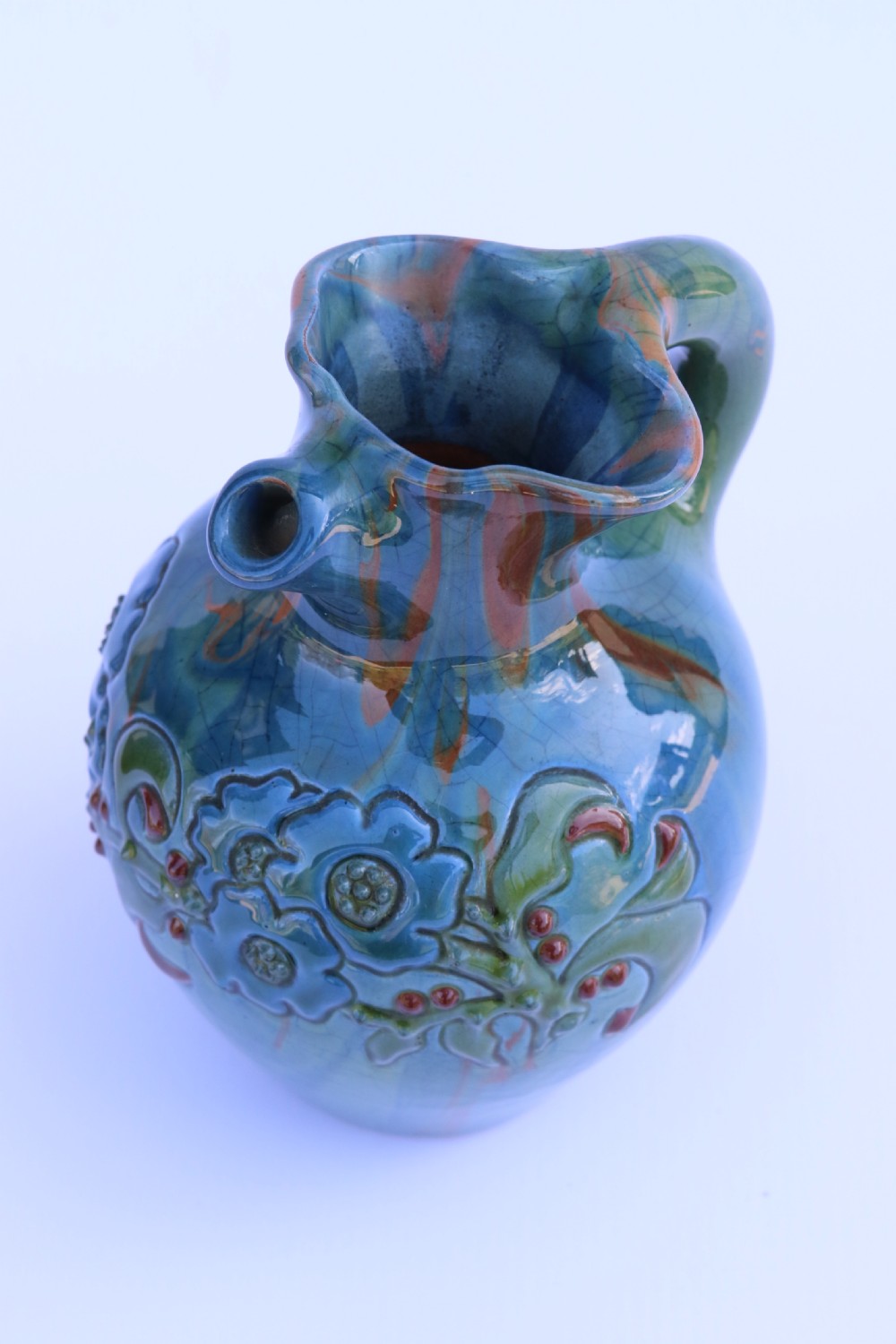A teal blue Arts and Crafts earthenware studio pottery jug featuring raised floral slipwork motifs, red glazed highlights, and a pinched spout. View XII