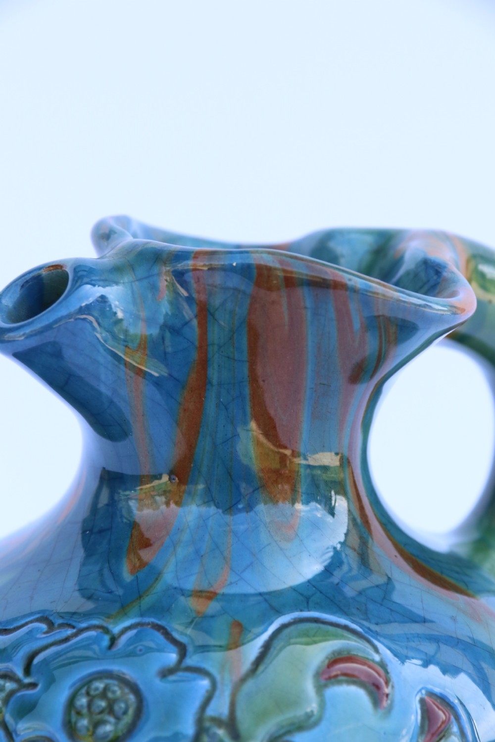 A teal blue Arts and Crafts earthenware studio pottery jug featuring raised floral slipwork motifs, red glazed highlights, and a pinched spout. View XIV