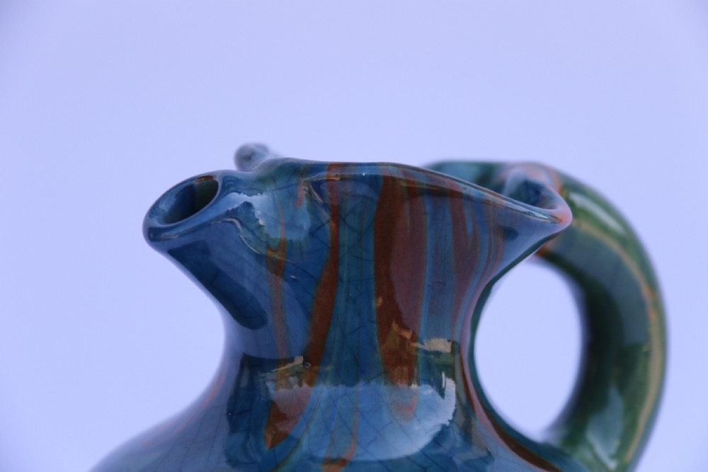 A teal blue Arts and Crafts earthenware studio pottery jug featuring raised floral slipwork motifs, red glazed highlights, and a pinched spout. View XV