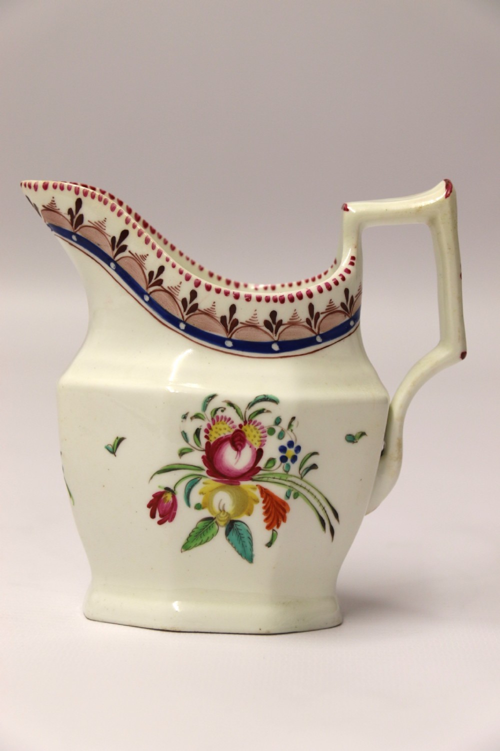 An 18th-century cream-coloured porcelain jug featuring hand-painted floral motifs, a decorated rim border, and an angular handle.