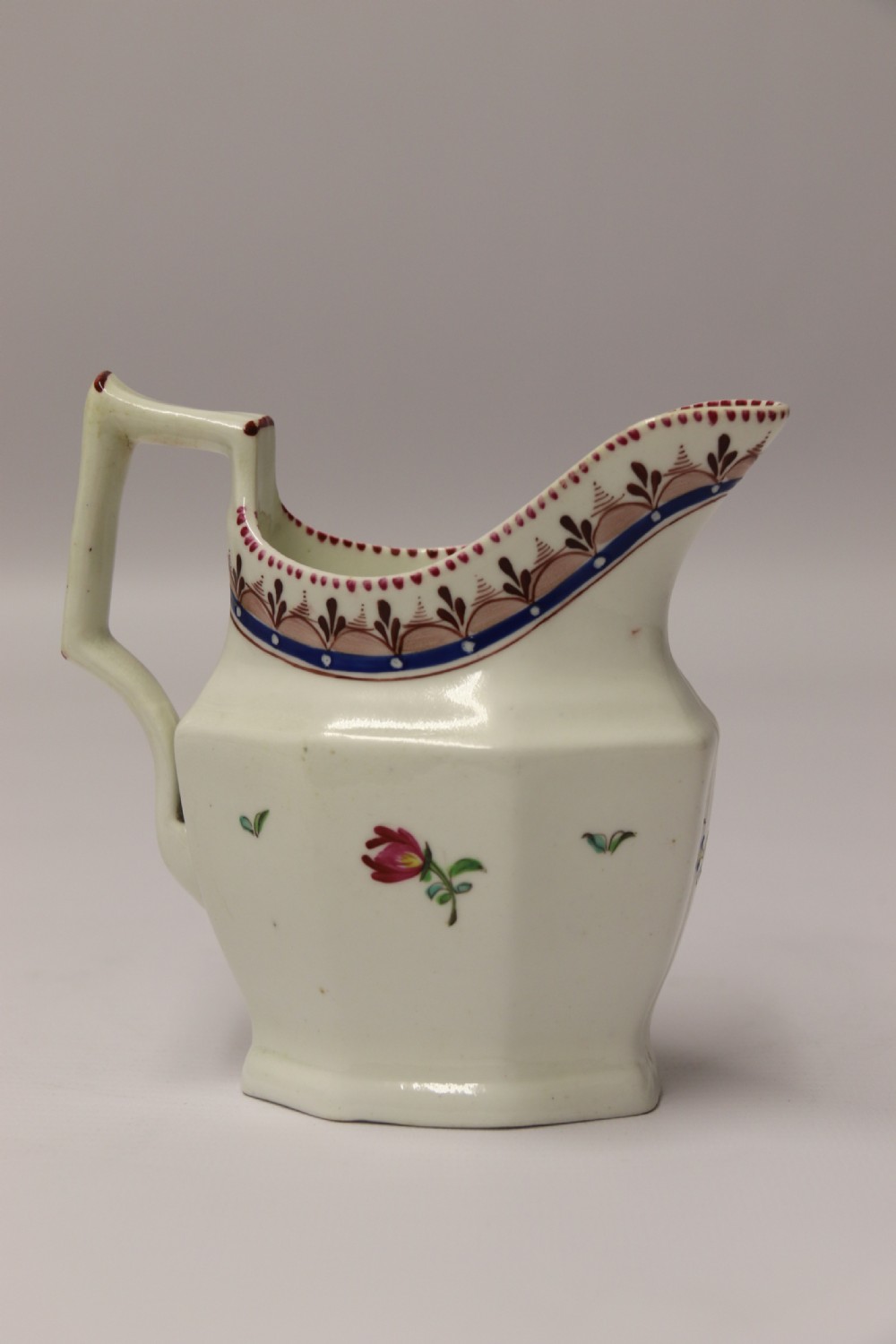 An 18th-century cream-coloured porcelain jug featuring hand-painted floral motifs, a decorated rim border, and an angular handle. View V