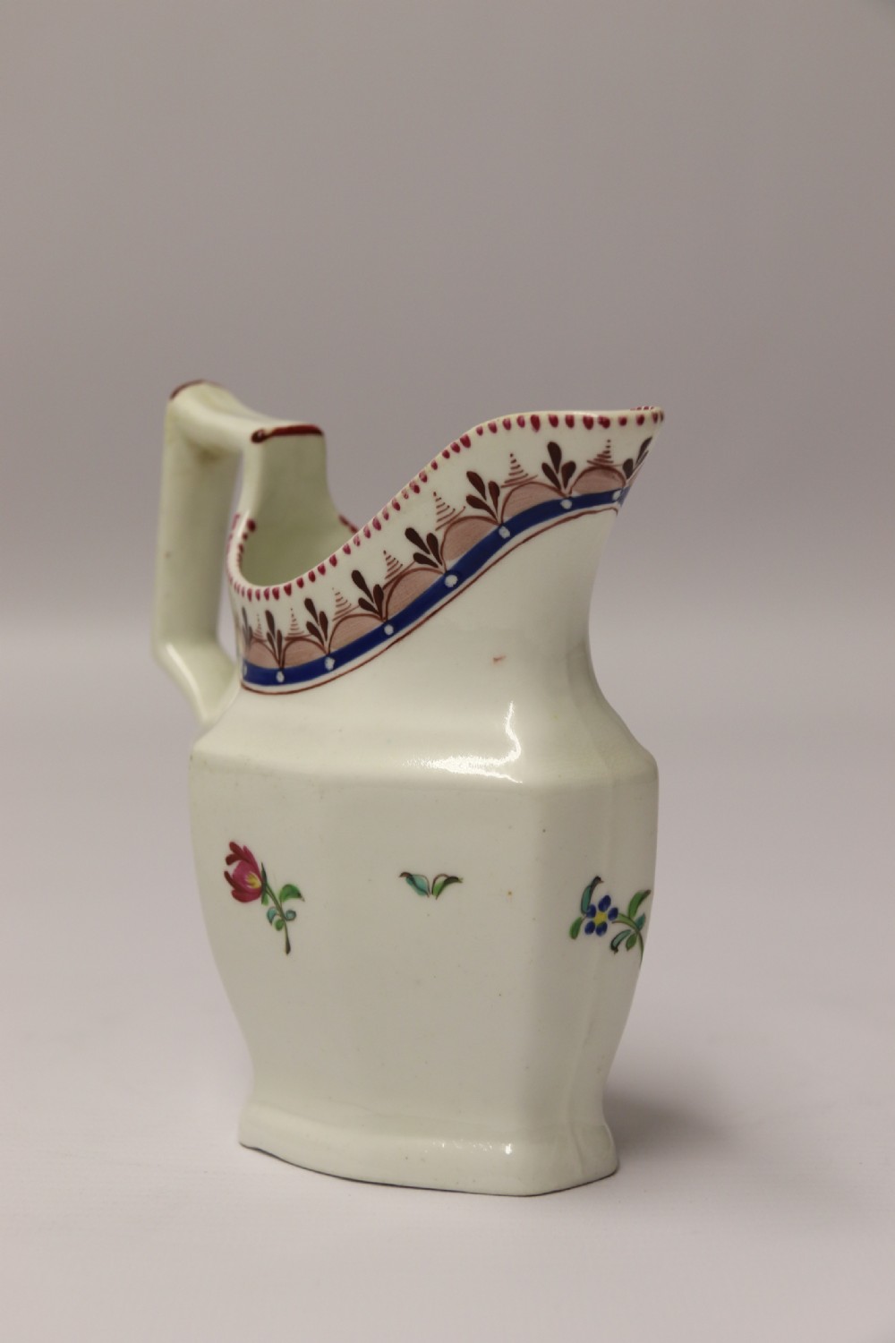 An 18th-century cream-coloured porcelain jug featuring hand-painted floral motifs, a decorated rim border, and an angular handle. View VI