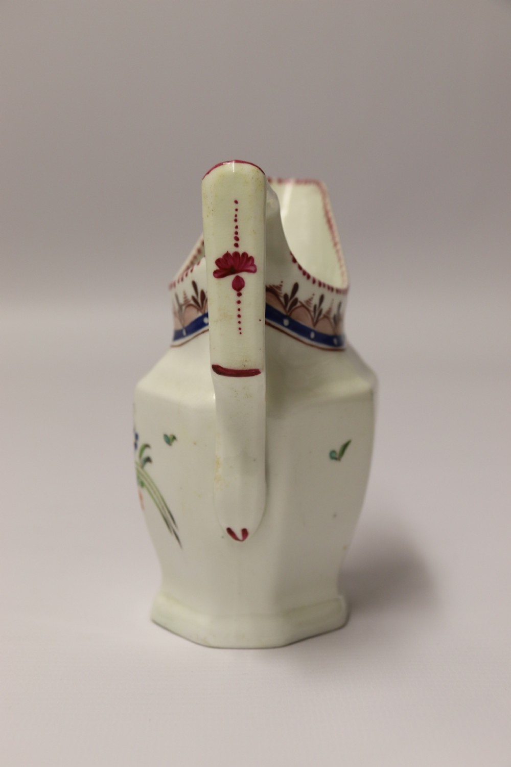 An 18th-century cream-coloured porcelain jug featuring hand-painted floral motifs, a decorated rim border, and an angular handle. View VIII