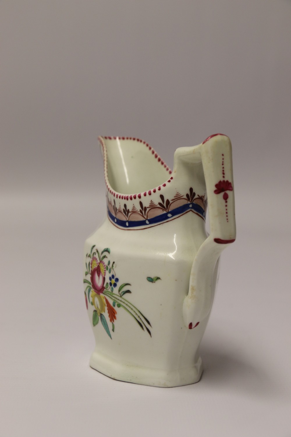 An 18th-century cream-coloured porcelain jug featuring hand-painted floral motifs, a decorated rim border, and an angular handle. View IX