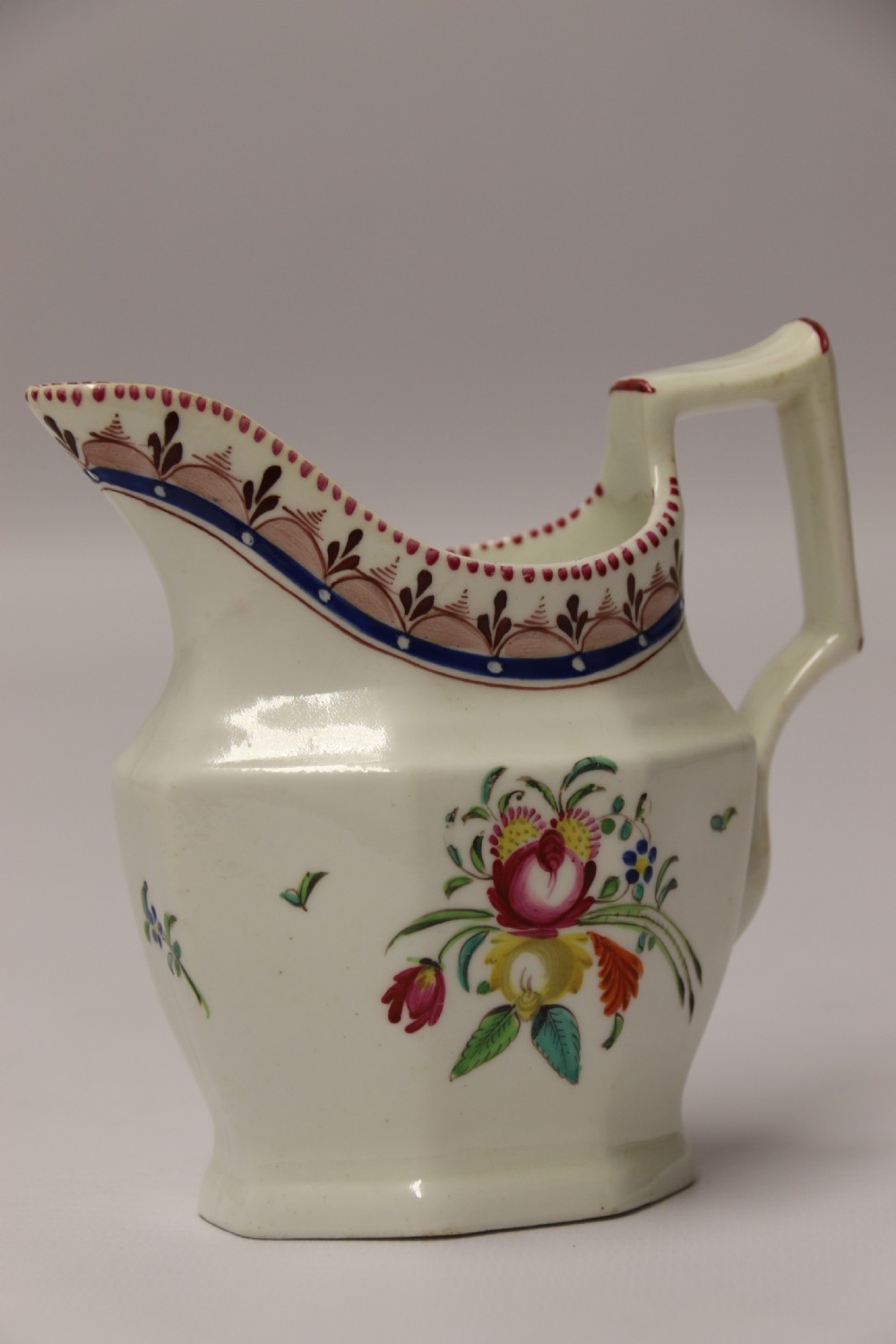 An 18th-century cream-coloured porcelain jug featuring hand-painted floral motifs, a decorated rim border, and an angular handle. View XII